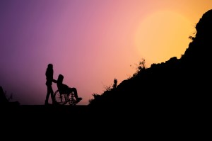 Individual pushing another person in a wheelchair with sunset.